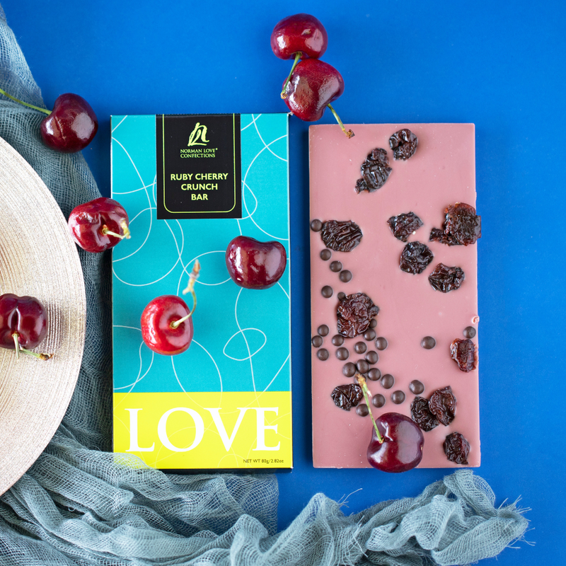 A pink ruby cherry crunch chocolate bar, studded with chocolate pearls and dried cherries, next to a teal chocolate bar wrapper with cherries scattered throughout.