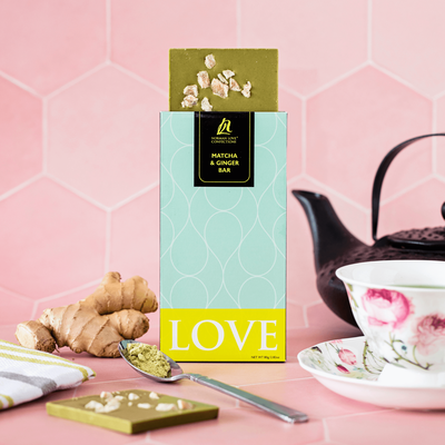 Piece of ginger; square of green matcha ginger chocolate bar coming out of teal package; black tea kettle (background); green napkin; spoon with matcha; rose tea cup & saucer (middleground); green chocolate bar (foreground) on pink background.