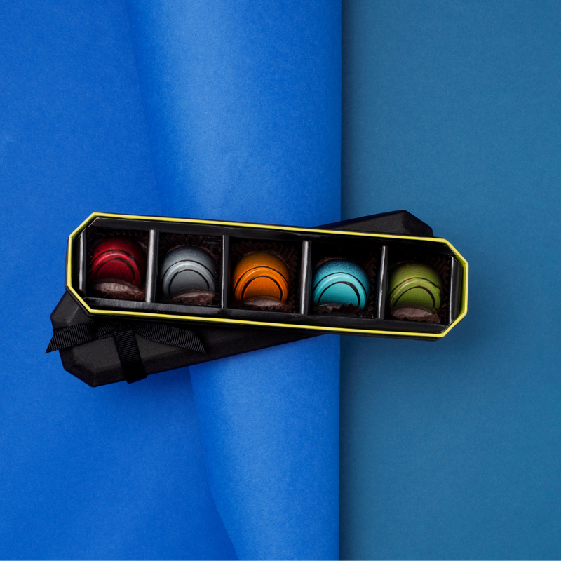 5 piece "BLACK" dark chocolates on black lid. Chocolates are red, grey, orange, blue and green. Black lid below open box.