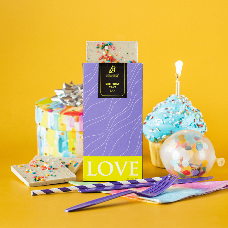A purple & green chocolate bar package, with a birthday cake bar emerging from the top. Pieces of the chocolate bar are scattered showing its white color and sprinkle topping. A cupcake with a candle and party supplies in the background evoke the flavor.