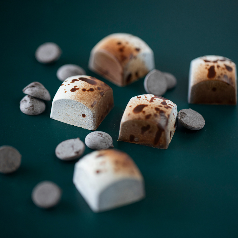 5 Tiramisu chocolates on a green background with loose coffee beans scattered across. The bonbons are square with a rounded top, and feature a half white, half brown design speckled with dark brown throughout.