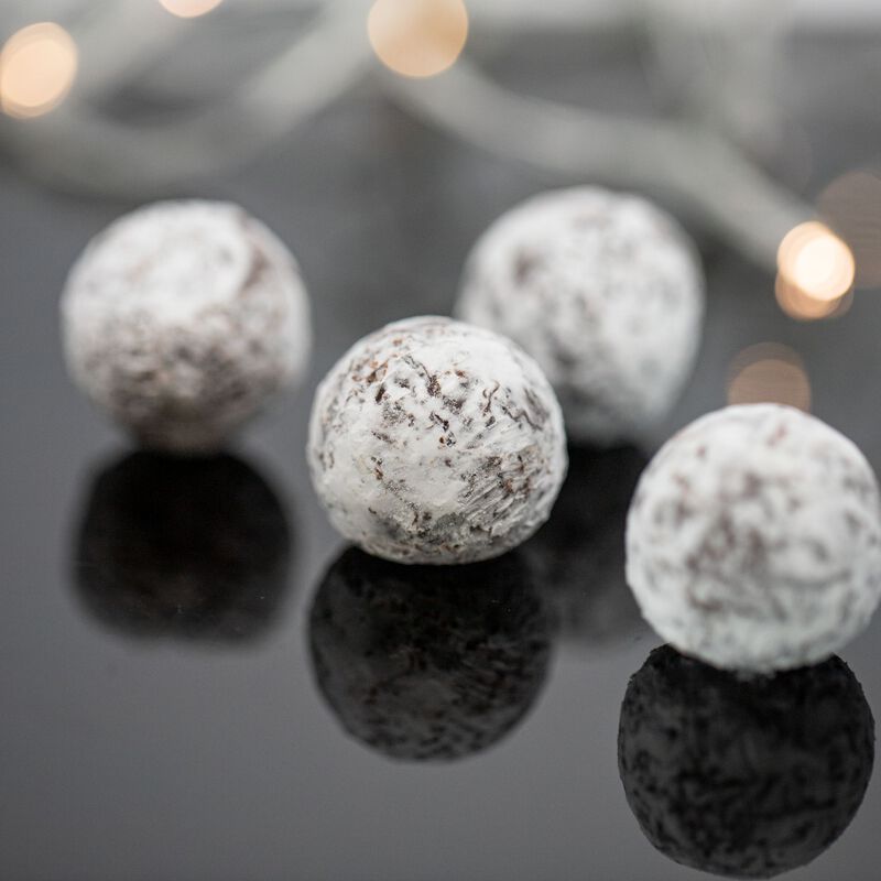 Four round, white champagne truffles on a reflective glass surface, with white Christmas lights in the background. The truffles are round and show glimpses of brown beneath a powdery white coating.