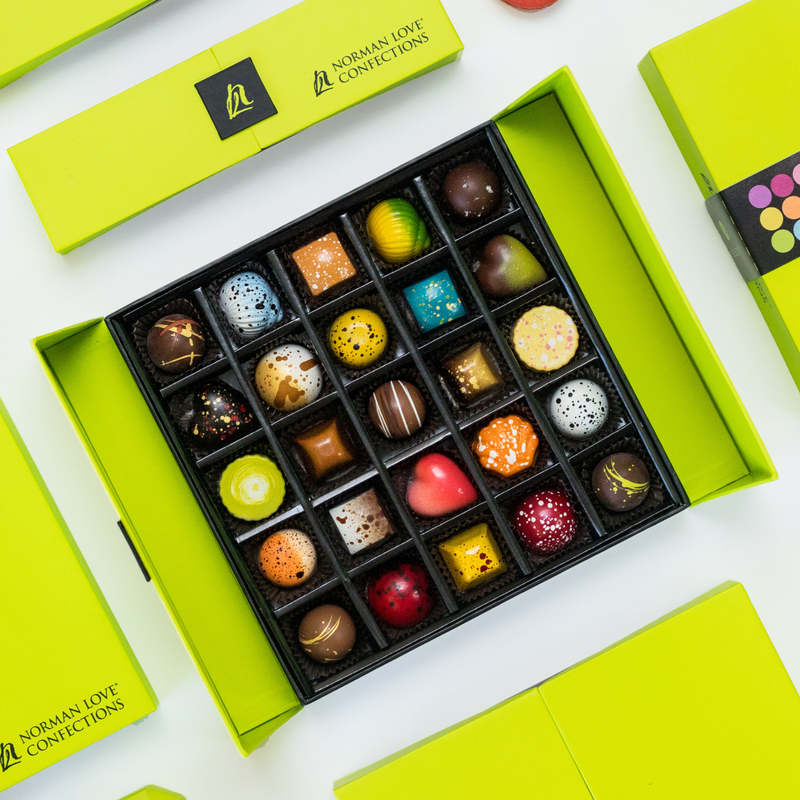 An overhead view of one open 25-Piece Signature Gift Box tilted at an angle and bordered by other stylish gift boxes. The open box shows an array of brightly colored chocolates in different artistic shapes. 