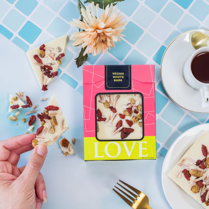 Vegan white chocolate bar in pink box on blue tile background. Flowers(left)and a coffee cup(right).