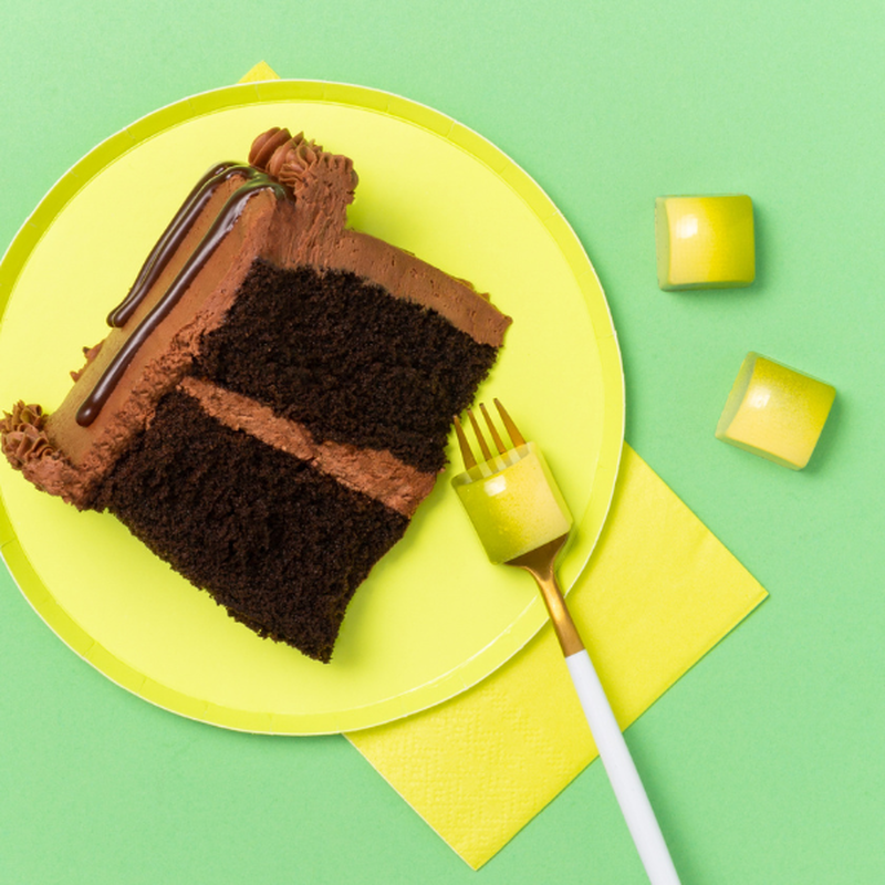 A slice of chocolate cake on a plate with a fork and three green & yellow pieces of German Chocolate Cake chocolates scattered loosely to evoke the flavor.