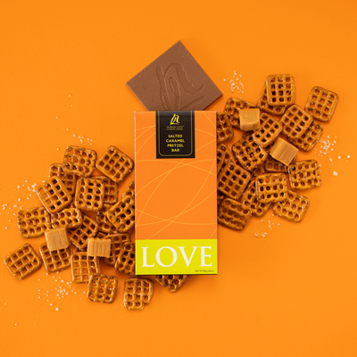 Orange pretzel chocolate bar on orange background. Checkerboard pretzels, square caramels, and sea salt scattered throughout. Square chocolate bar shown coming out of packaging. 
