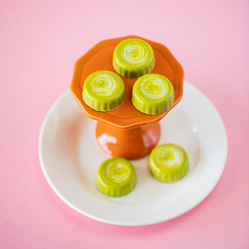 5 Peanut Butter Cup Chocolates, 3 sitting atop an orange pedestal which sits on a white plate with the other 2 pieces. Shot at an angle to show the scalloped edge & green color of the chocolate.