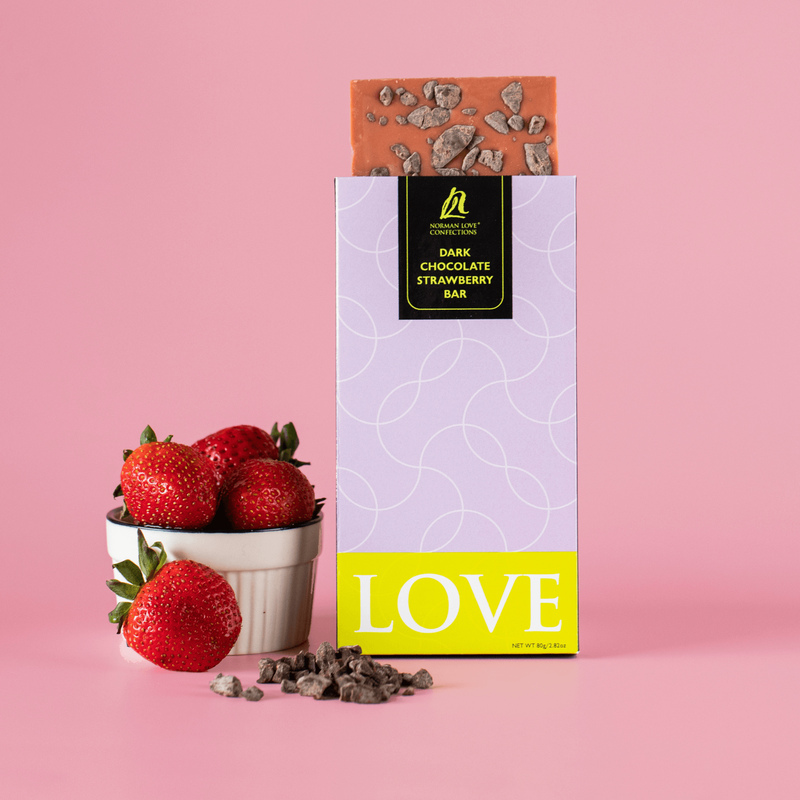 White bowl of red strawberries (left) dark chocolate strawberry bar in light purple packaging (center) with unwrapped pink bar at top, cacao nibs scattered in front of bar on pink background.