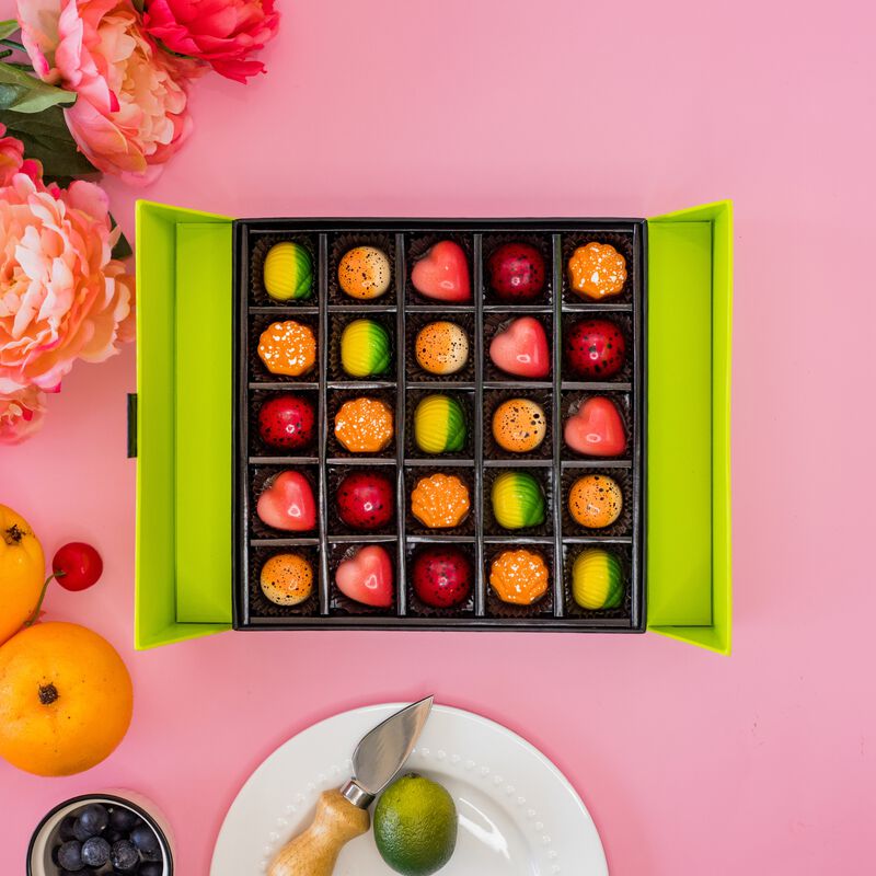 Open Fruit Favorites Gift Box displays brightly colored chocolates shaped as hearts, seashells, and circles. The 25 piece box sits on a pink background with pink flowers, oranges, a cherry, blueberries and a lime to represent truffle flavors. 