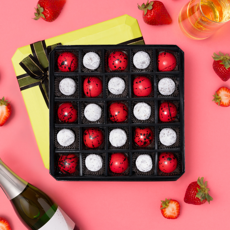 25 piece gift box of red & white bonbons circular bonbons. Green lid with brown bow underneath open box. Strawberries, glass of champagne and champagne bottle on pink background.