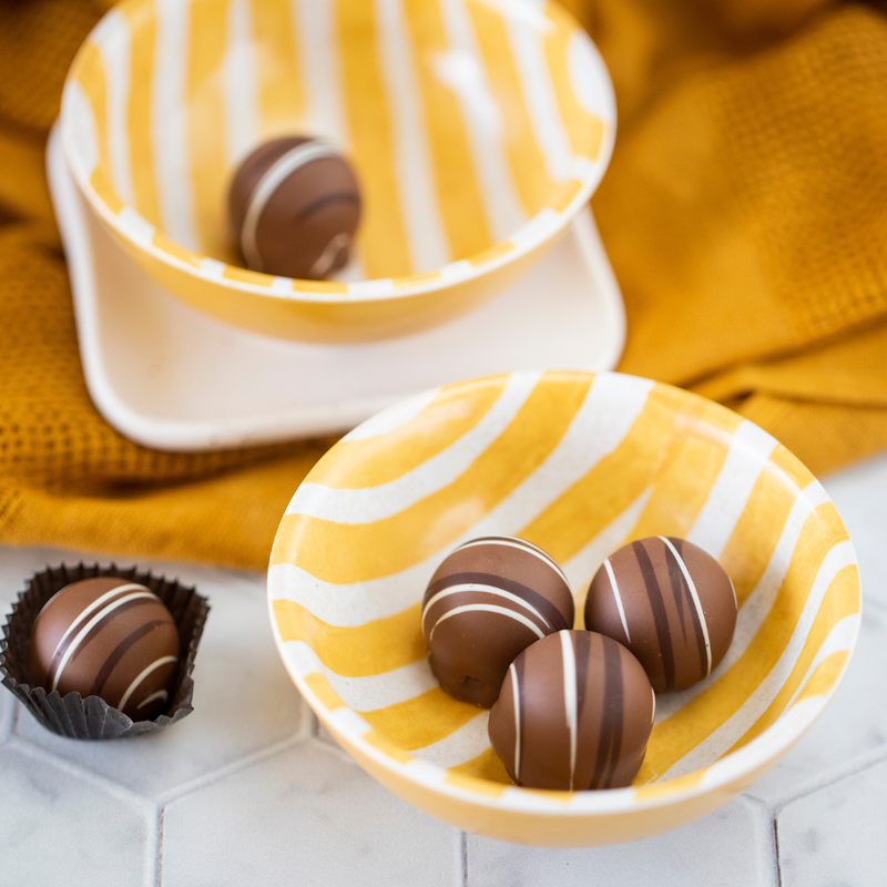 Five roasted almond truffles, 4 in yellow and white striped bowls and one sitting in a brown paper chocolate wrapper, arranged to show the circular truffles' white and dark brown stripes and light brown coloration.
