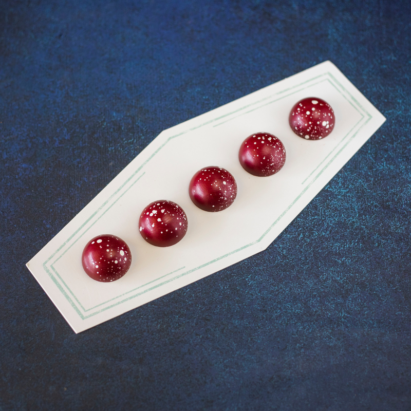 Overhead shot of 5 Raspberry chocolates on an oblate white plate over a blue background. The chocolates are red and dome-shaped.