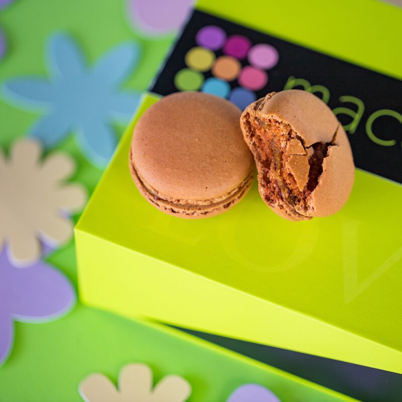Two Dark Chocolate French macarons atop a beautiful lime green gift box. One macaroon is bitten into to show the chewy center. Flower cut-out decorations provide texture to the green background. 
