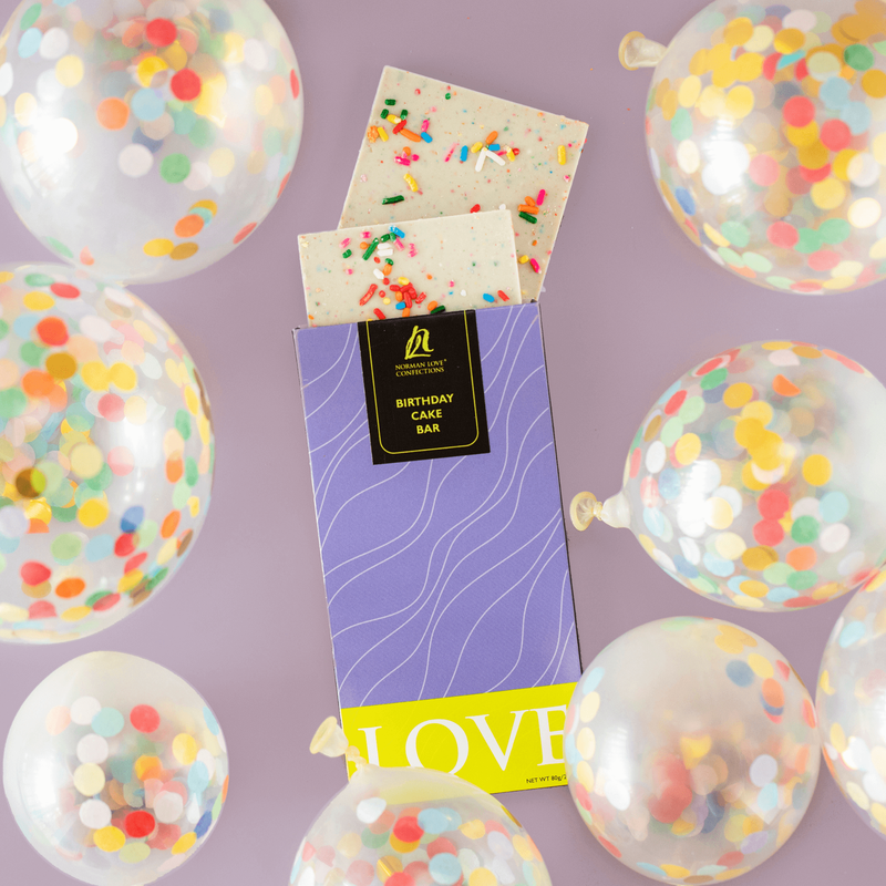 A purple and green chocolate bar package, with two pieces of white chocolate bar poking out the top, showing its white color and sprinkle topping. Multicolored party balloons surround the chocolate bar to evoke the flavor.