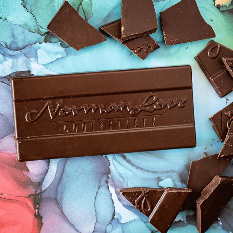 A Dark Chocolate Bar lying flat on blue marbled background, with pieces of dark chocolate bar scattered behind it. The bar is dark brown and emblazoned with the Norman Love Confections logo.