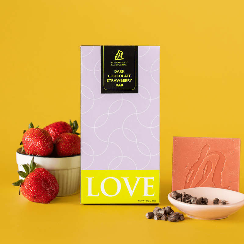 White bowl of red strawberries and a dark chocolate strawberry bar in light purple packaging, featuring an unwrapped pink chocolate bar behind dish of cacao nibs (right) on yellow background. 