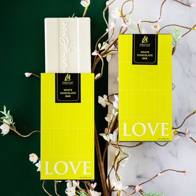 White Chocolate Bar coming out of lime green packaging on dark green background to the left. White chocolate bar in lime green packaging on white marbled background to the right. White foliage behind both chocolate bars. 