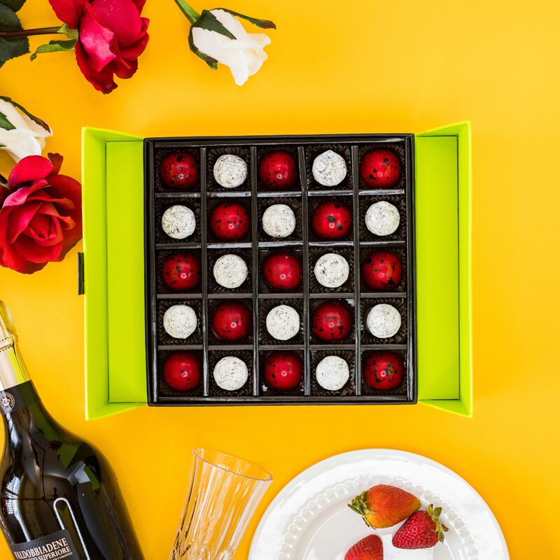 Open 25 piece champagne and strawberries gift box. Chocolates are red and white in a green box. White and red flowers on an orange background. 