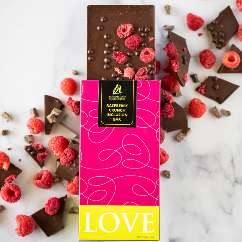 Pink packaged raspberry crunch inclusion bar on white marbled background. Dark chocolate bar with pink raspberries coming out of top of package. Raspberries and chocolate pieces scattered diagonally (left to right) behind bar.