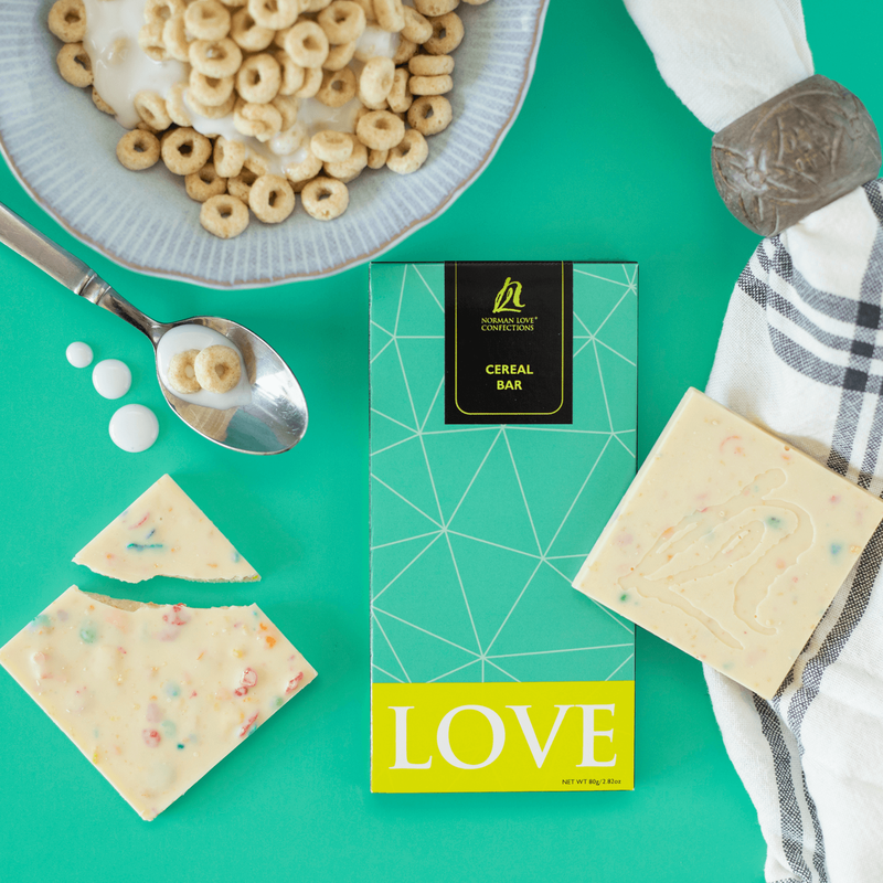 Teal wrapped cereal bar in center of teal background. Light blue bowl of cereal and milk, spoon with cereal and milk, white cereal bar with multi-colored cereal to left. White and black napkin with wood napkin ring and unwrapped cereal bar on right. 
