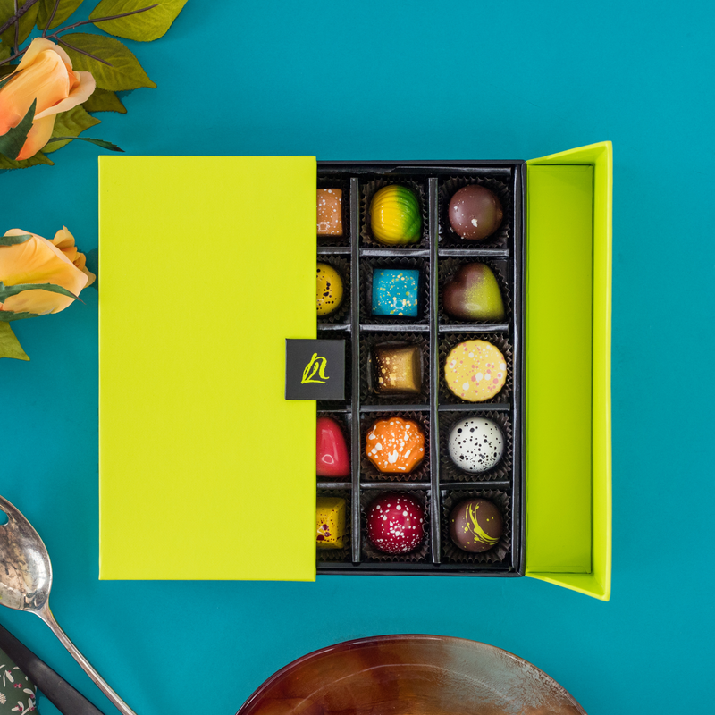 A half open 25-PIece Signature Gift Box displays a variety of artistically crafted signature chocolates. The brightly colored chocolates pop against a dark teal background. Soft orange roses, a silver spoon and a brown plate surround the gift box.