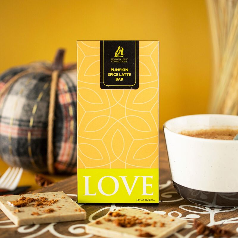 Rectangle Pumpkin Spice Latte bar shown next to white coffee mug. Wrapping is orange & white, neon green "LOVE" label (bottom).  Pieces of bar with pumpkin spice scattered on top (foreground). Black, white, red, gold pumpkin and straw (background).