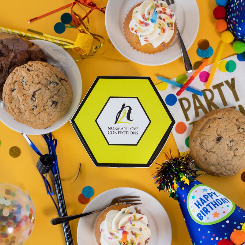 Chocolate chip & chocolate chunk cookie, 2 white cupcakes with white frosting and sprinkles surround hexagon, lime green cookie box. Confetti blue party hats and multi-colored candles fill scene. 
