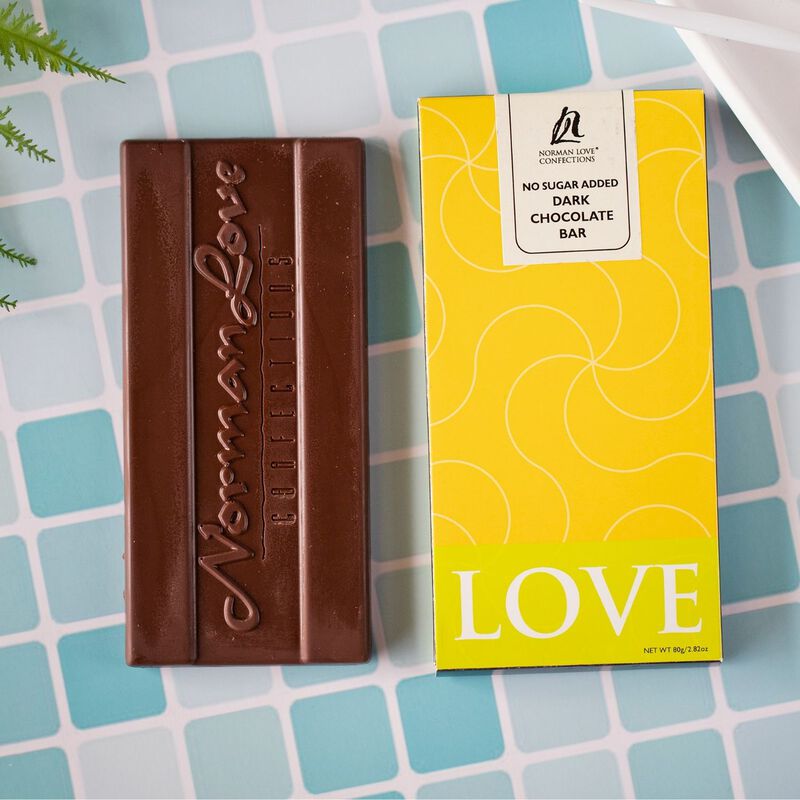 Wrapped and unwrapped No Sugar Added Dark Chocolate Bar on blue tiled background. Wrapped bar is in yellow packaging, unwrapped bar has "Norman Love Confections" printed on it.  