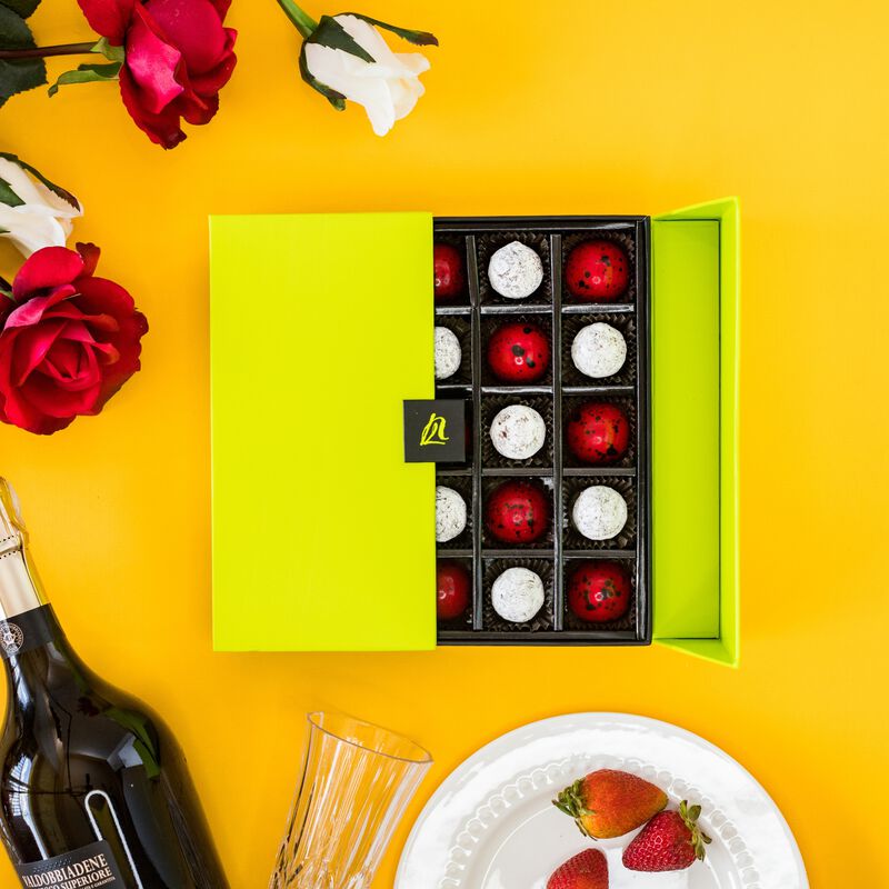 Half open 25 piece champagne and strawberries gift box. Chocolates are red and white in a green box. White and red flowers on an orange background. 