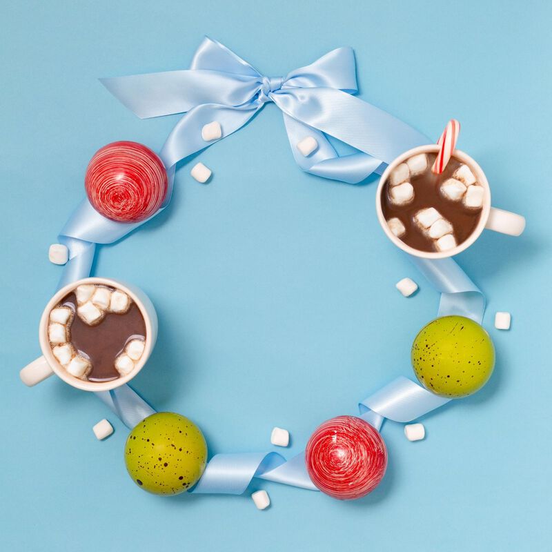 Four hot chocolate bombs, placed on a decorative blue ribbon to form a wreath. Features two signature green hot chocolate bombs and two red peppermint hot chocolate bombs and two cups of marshmallow-filled hot cocoa to bring the concept to life.