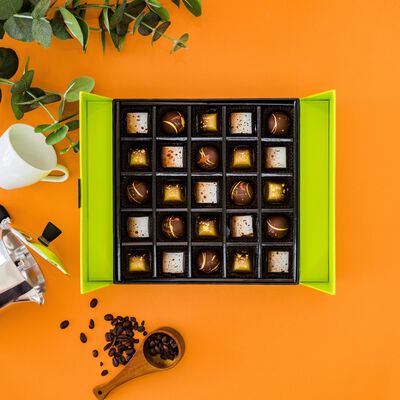 Open coffee shop gift box on an orange background. Chocolates are white and gold circles and squares. Greenery, white coffee cup, french press and coffee beans on a wooden spoon surround gift box. 
