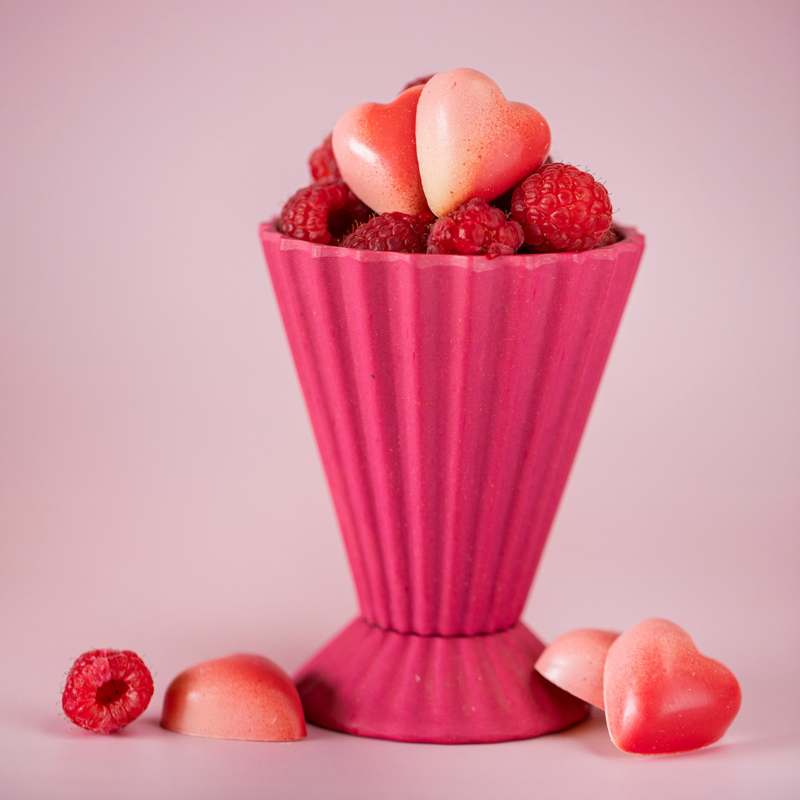 5 heart-shaped White Chocolate Raspberry chocolates. Two sit in a pink vase atop a layer of raspberries, while the other three sit at its base, arranged to show the chocolates' two-tone pink color.