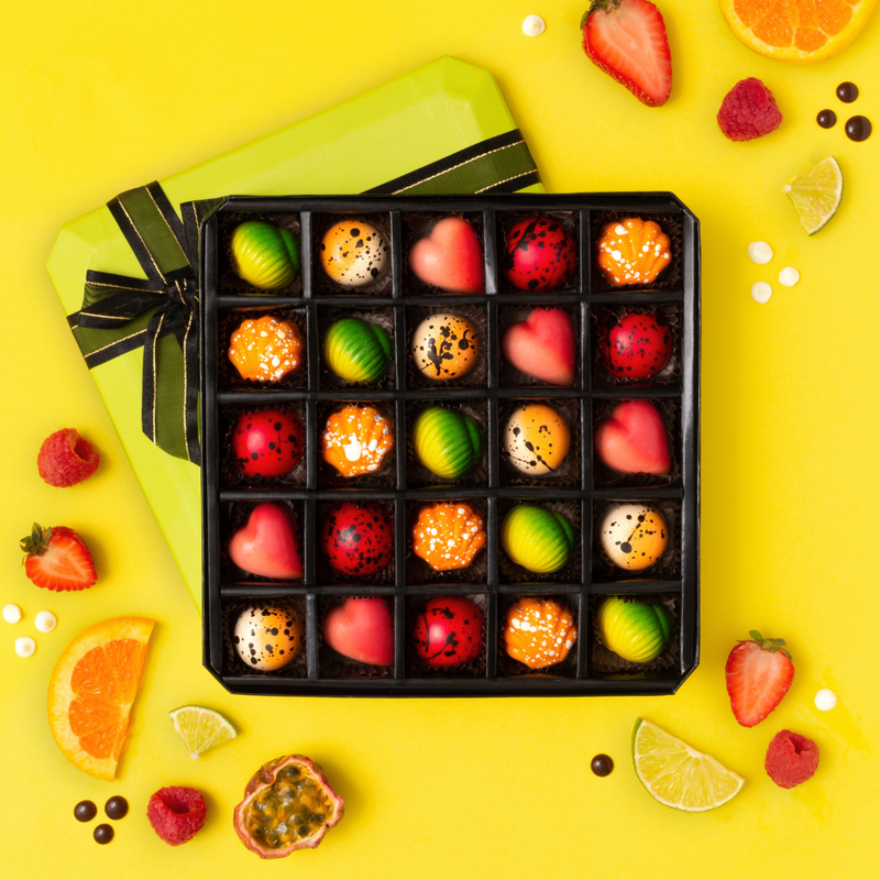 25 piece box of red, pink, yellow, green and orange bonbons on yellow background. Bonbons are hearts, seashells, and circles. Green lid with brown bow underneath box. Pieces of fruit scattered around box. 