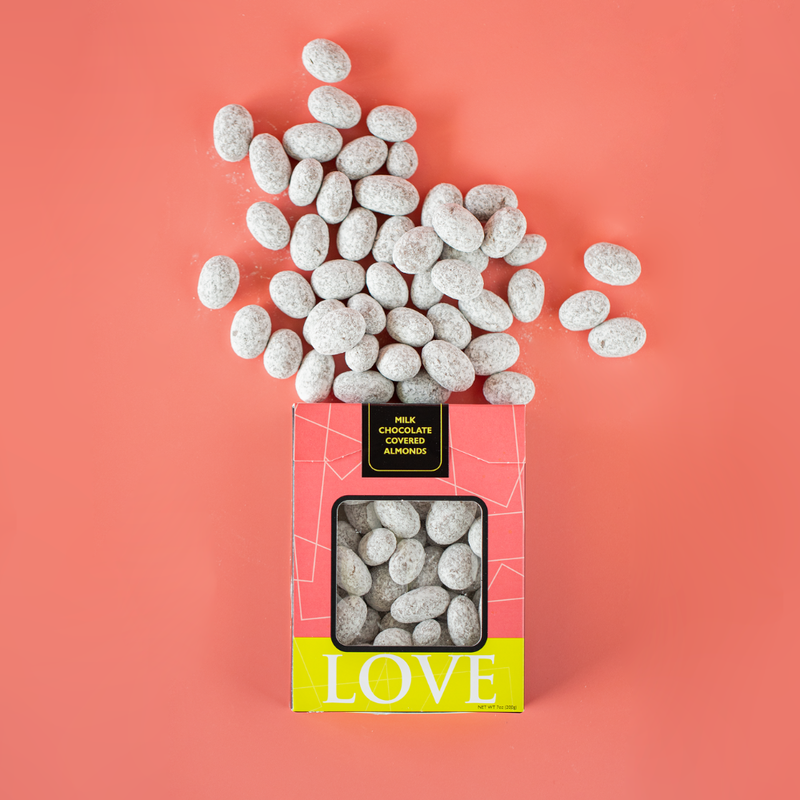 A pink box of milk chocolate covered almonds, with white powdered sugar covered almonds scattered around on a pink background.