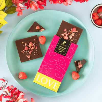 Dark chocolate raspberry crunch bar in pink packaging tilted on blue plate. Dark chocolate bar with raspberries coming out of top of package. Square bar, broken pieces & raspberries scattered on plate. Flowers and bowl of raspberries surround plate. 