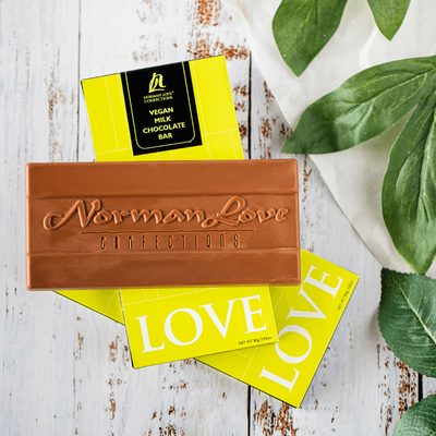 Unwrapped Vegan Milk Chocolate bar with "Norman Love Confections" printed in chocolate, stacked on two lime green wrapped bars, on white wood background. Green plant leaves on white napkin to the right.