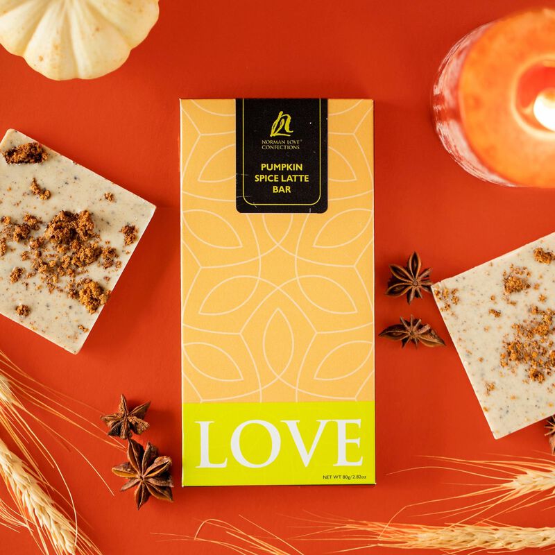 Rectangle wrapped Pumpkin Spice Latte bar in the center of orange background. Wrapping is orange & white, neon green "LOVE" label. White pumpkin, star anise pods, orange candle, unwrapped white bar with pumpkin spice seasoning around bar. 
