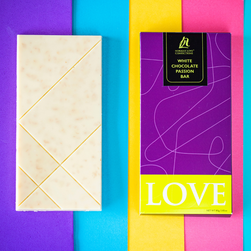 A white chocolate passionfruit bar, featuring pieces of passionfruit set in a white bar of chocolate, next to a purple chocolate bar wrapper on a blue, purple, yellow and pink background.