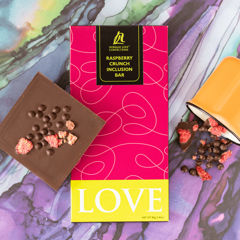 Dark Chocolate Raspberry Crunch Bar (center) in pink packaging multi-colored marble background. Square bar with raspberries and chocolate pearls (left); tilted orange mug of raspberries and chocolate pearls (right). 