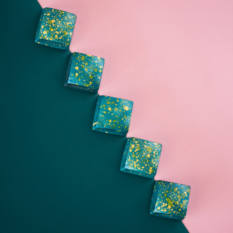 Overhead view of 5 blue 80% Coeur de Guanaja bonbons, arranged in a diagonal on a background of green and pink, showing the square shape and green and yellow speckles on each piece.