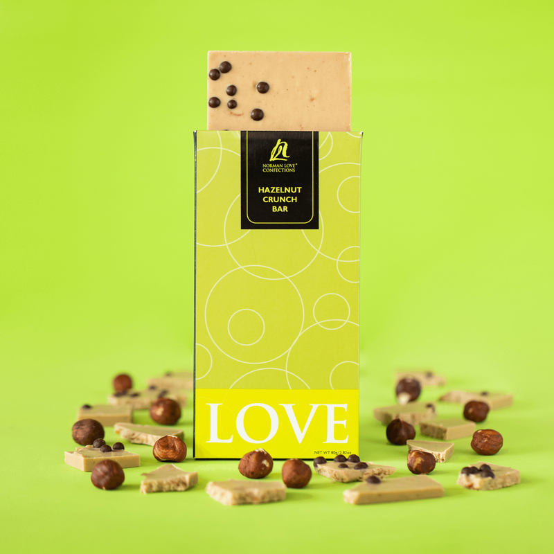 Hazelnut Crunch Bar (center) in lime green packaging on lime green background. Square chocolate bar is coming out of package with hazelnuts and chocolate pieces scattered around bar. 