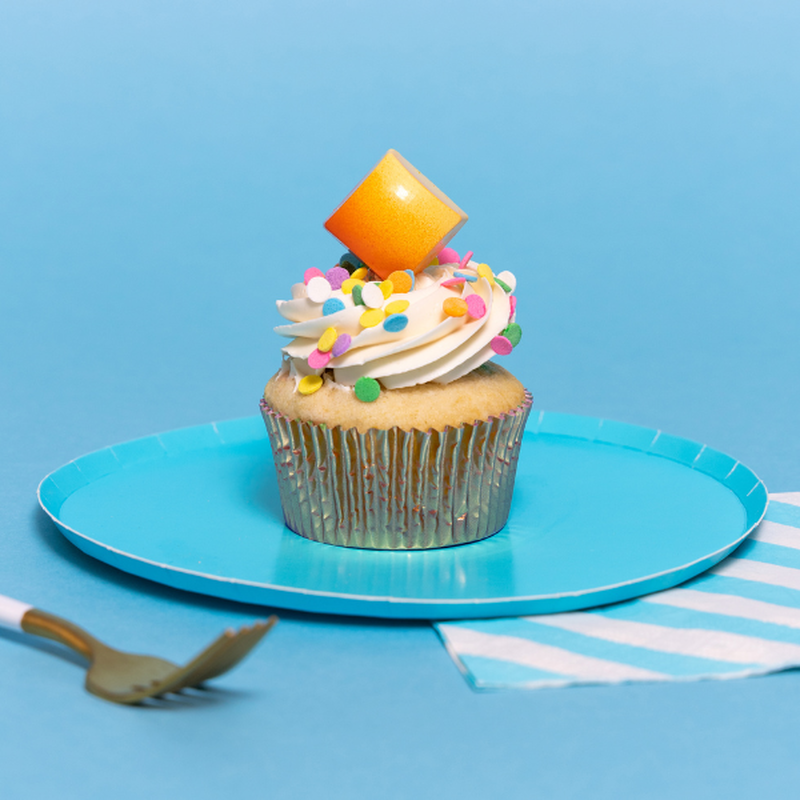 A blue plate with a confetti-adorned cupcake on it. On top of the cupcake is a Cupcake chocolate, angled into the frosting to show its orange & yellow patterning and square shape with a fork in the periphery.