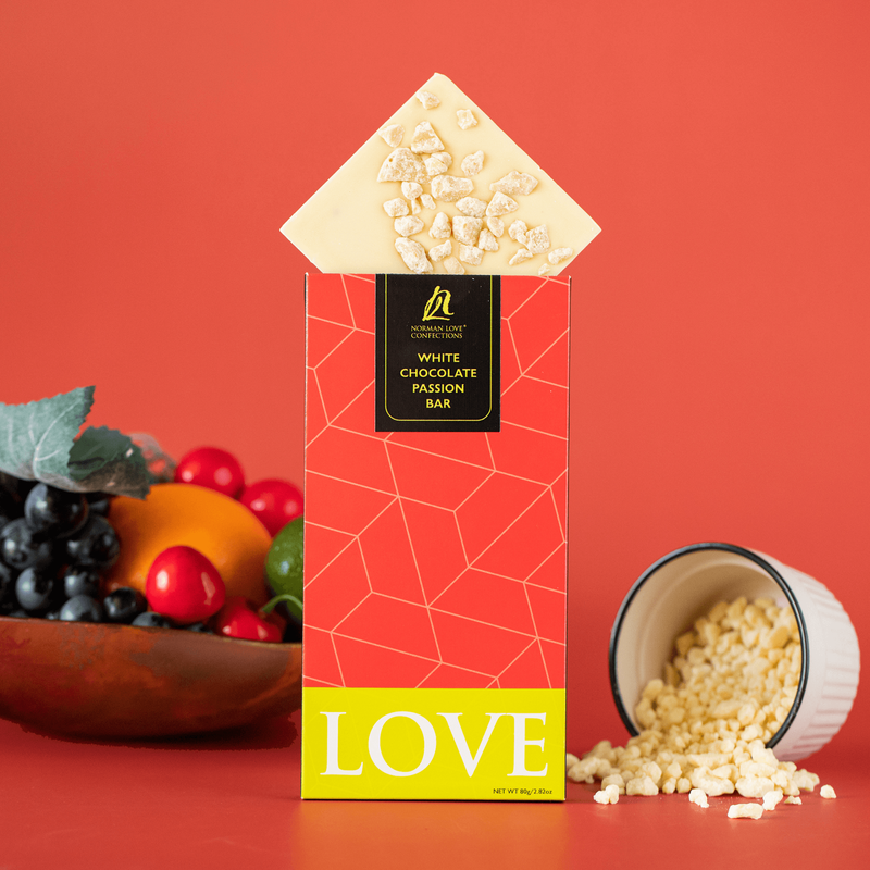 Wood bowl of fruit; tilted square piece of white chocolate passion bar on top of red rectangle packaged bar (center); white chocolate falling out of tipped white bowl with black rim (right) on red background. 