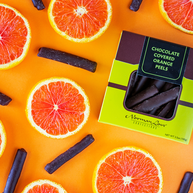 Lime green box of chocolate covered orange peels on orange background. Open oranges and pieces of dark chocolate orange peels scattered throughout. 