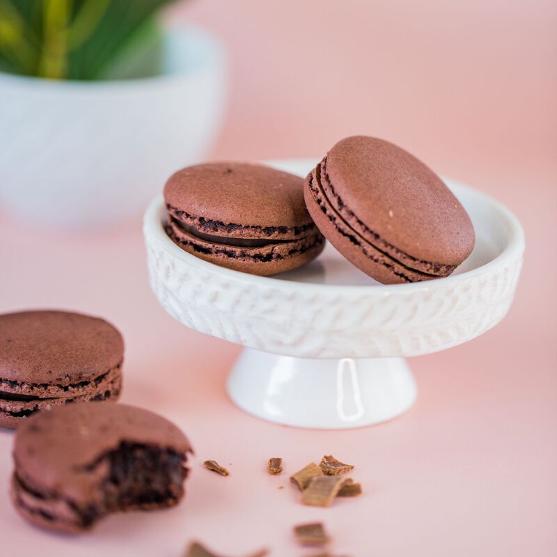 Four Dark Chocolate French Macaroons displayed on a rose-pink background. Two are on a white dessert stand and two are beside the stand with chocolate shavings. Each macaron displays a ruffled edge and gooey center to show the quality of the cookie.
