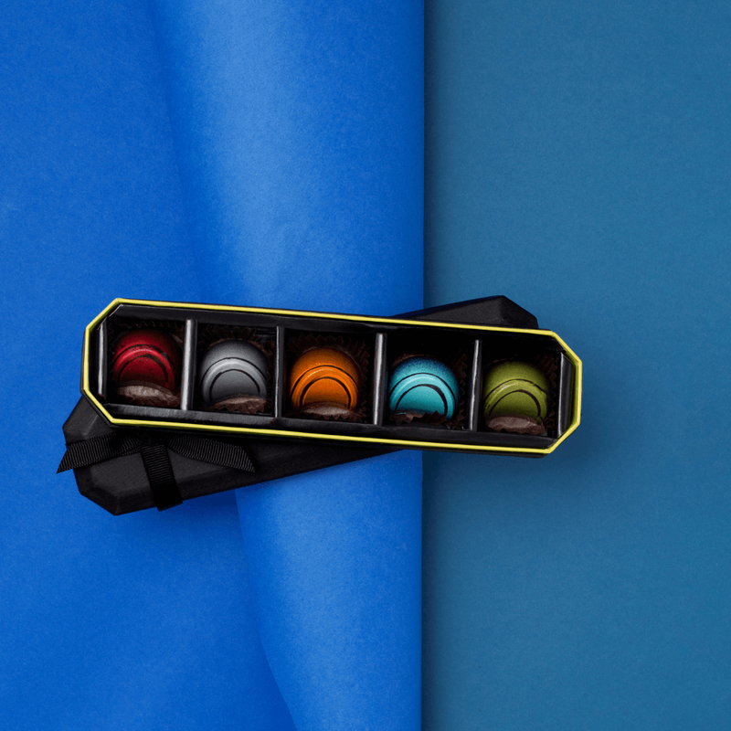 5 Piece BLACK Dark Chocolate Gift Box, opened to show five different shapes, colors, and varieties of chocolates on a blue background. Flavors: Jiquilisco Bay (97%), Maracaibo (88%), Tanzanie (75%), Pico Bonito (70%), Nyangbo (68%).