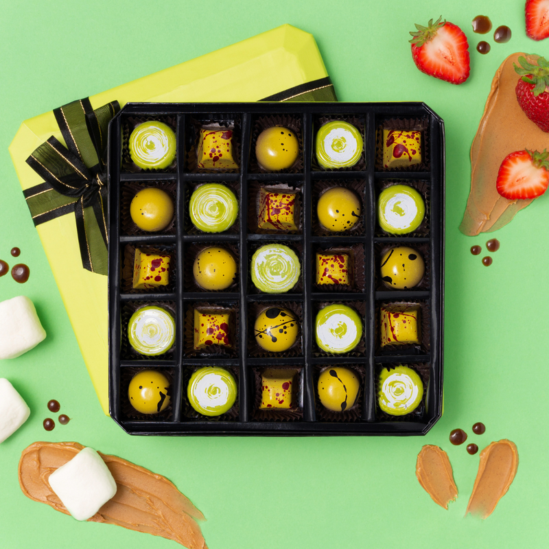 25 piece box of yellow and green chocolates on green background. Lime green box lid with brown bow below open chocolate box. Peanut butter smudges, marshmallows and strawberries surround box. 