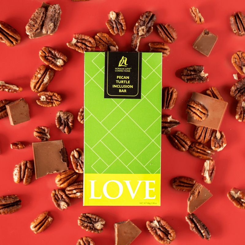Pecan Turtle Bar (center) in bright green packaging on red background. Pieces of pecans and chocolate scattered around. 