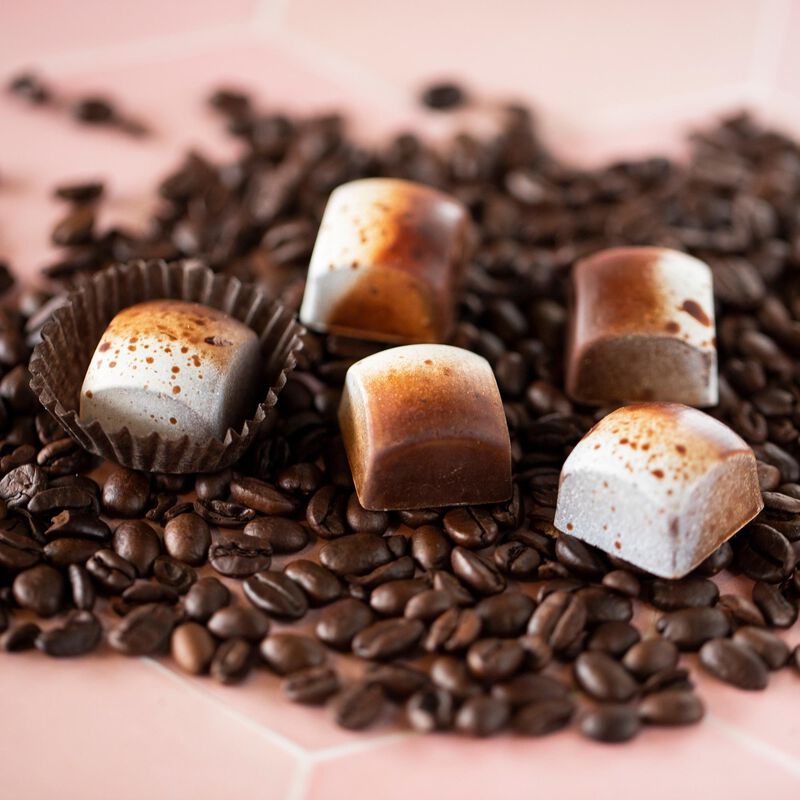 4 Tiramisu chocolates sitting on a pile of coffee beans and 1 Tiramisu chocolate sitting in a paper cup. The chocolates are square and arranged to showcase their brown and white coloration, which is flecked with light brown spots.