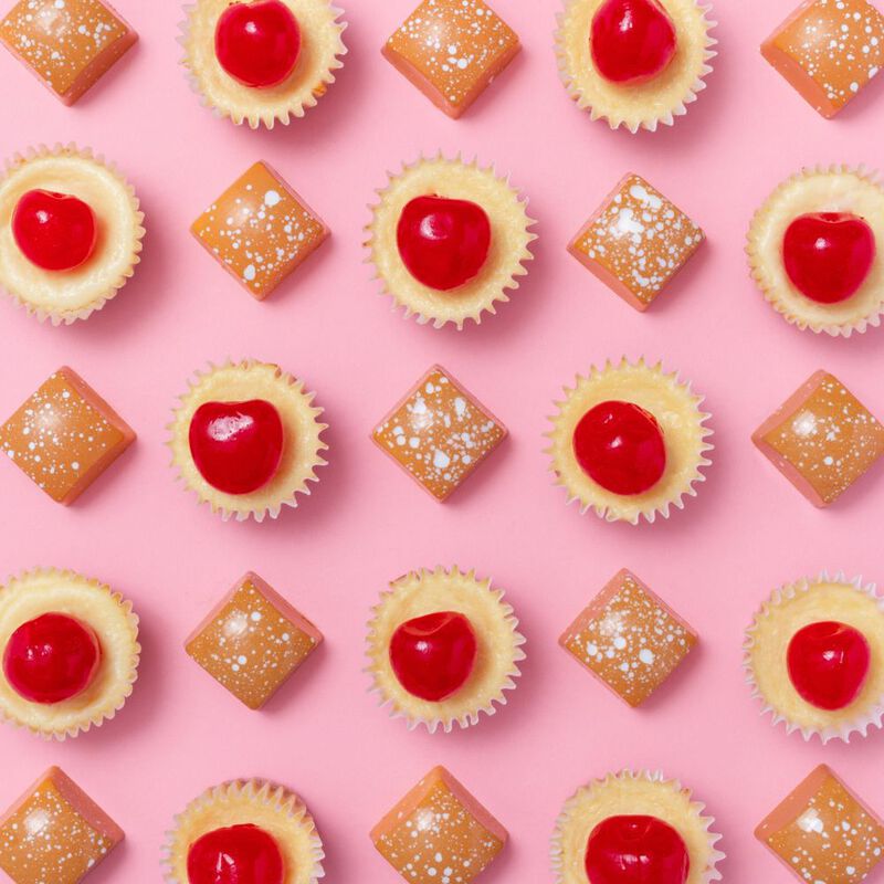 Tan, speckled New York Cheesecake chocolates and mini cupcakes topped with cherries, arranged in a grid alternating between chocolates and cupcakes